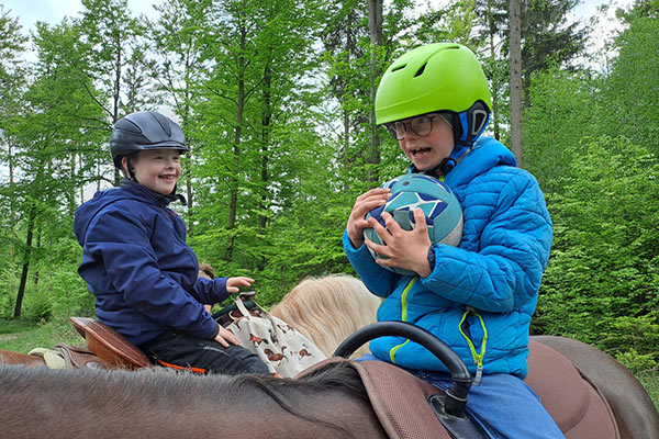 Zwei Kinder auf zwei Pferde, die im Rahmen der Pferdegestützten Therapie mit einem Ball spielen.